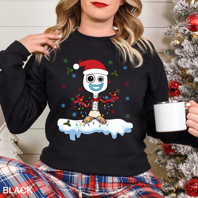 Puede incluir: Sudadera negra con un personaje de dibujos animados que se asemeja a un tenedor-cuchara con un gorro de Pap&aacute; Noel y luces navide&ntilde;as. El dise&ntilde;o incluye copos de nieve y acebo. La palabra "BLACK" est&aacute; impresa en la parte inferior. Una taza blanca se sostiene en la mano.