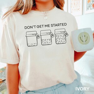 May include: Ivory-colored t-shirt with the text "DON'T GET ME STARTED" above three jars, each with a tag. The jars are filled with varying amounts of dots. A white mug with a smiley face is on the right.