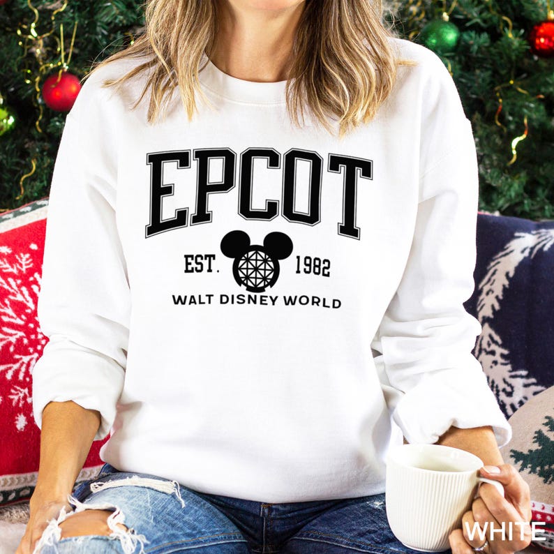 K&ouml;nnte beinhalten: Wei&szlig;es Sweatshirt mit dem Wort "EPCOT" in schwarzen Blockbuchstaben. Darunter ein Mickey Mouse-Kopf mit Globe-Design, der Text "EST. 1982" und "WALT DISNEY WORLD". Das Sweatshirt hat einen Rundhalsausschnitt.