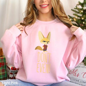May include: Pale pink sweatshirt with a cartoon fox illustration. The fox has an angry expression and is wearing a maroon vest. The words "DON'T EVEN!" are printed in yellow. The sweatshirt is a crew neck style.