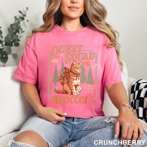 May include: A pink t-shirt with a graphic featuring a cat wearing a backpack, the words "Quest Squad" and "Broccoli". The design includes trees, a sun, and the text "No one wants it, but it's good for you."