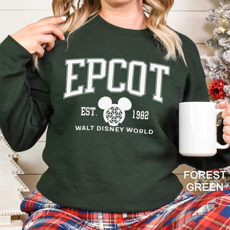 K&ouml;nnte beinhalten: Forstgr&uuml;nes Sweatshirt mit dem Wort "EPCOT" in wei&szlig;en Blockbuchstaben. Darunter steht der Text "EST. 1982" und "WALT DISNEY WORLD". In der Mitte befindet sich eine wei&szlig;e Mickey Mouse-Kopf-Grafik. Das Sweatshirt hat einen Rundhalsausschnitt.