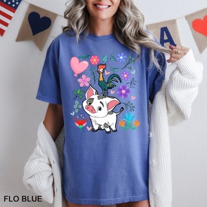 K&ouml;nnte beinhalten: Ein blaues T-Shirt mit einem Cartoon-Schwein und einem Hahn darauf. Das Schwein ist wei&szlig; mit schwarzen Flecken und der Hahn ist rot, gelb und gr&uuml;n. Um sie herum befinden sich Blumen und Herzen. Das Shirt tr&auml;gt am unteren Rand die Aufschrift "FLO BLUE".