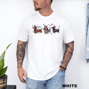 May include: White t-shirt featuring three dachshunds with patriotic bow ties and surrounded by blue stars. The dachshunds are in shades of brown, gray, and black. The word "WHITE" is printed in white at the bottom right.