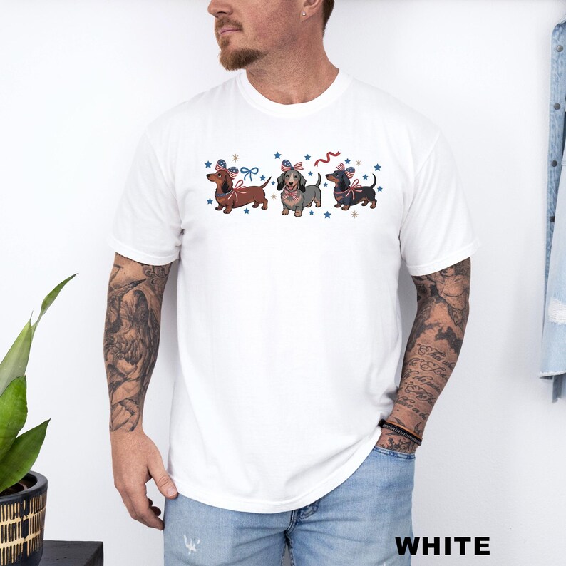 May include: White t-shirt featuring three dachshunds with patriotic bow ties and surrounded by blue stars. The dachshunds are in shades of brown, gray, and black. The word "WHITE" is printed in white at the bottom right.