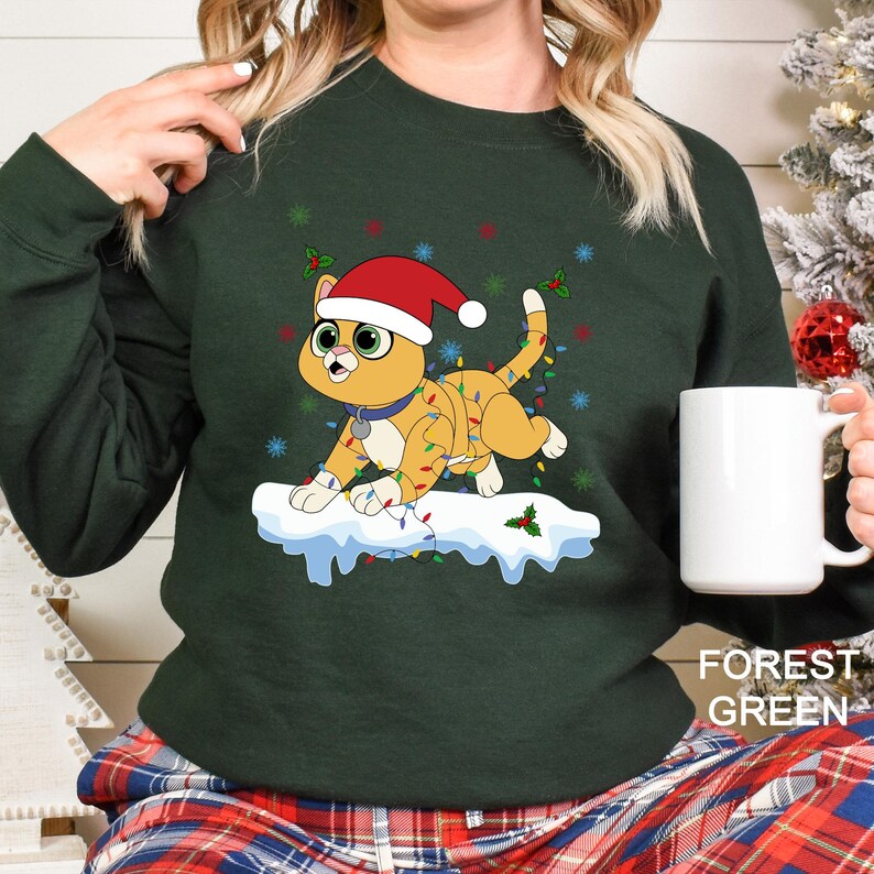 Puede incluir: Sudadera verde oscuro con un dise&ntilde;o festivo de gato de dibujos animados. El gato lleva un gorro de Pap&aacute; Noel y est&aacute; adornado con luces navide&ntilde;as, de pie sobre una base nevada. La sudadera tiene la frase "FOREST GREEN" impresa.