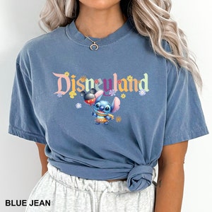 Puede incluir: Una camiseta azul con un gr&aacute;fico de Stitch de Lilo & Stitch sosteniendo un globo de Mickey Mouse. El texto "Disneyland" est&aacute; impreso sobre Stitch en una fuente colorida.