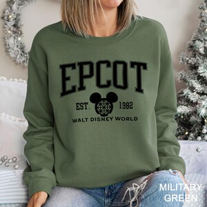 K&ouml;nnte beinhalten: Milit&auml;rgr&uuml;nes Sweatshirt mit dem Wort "EPCOT" in schwarzen Blockbuchstaben. Darunter ein Mickey Mouse-Kopf mit Globus-Design, der Text "EST. 1982" und "WALT DISNEY WORLD". Das Sweatshirt hat einen Rundhalsausschnitt.