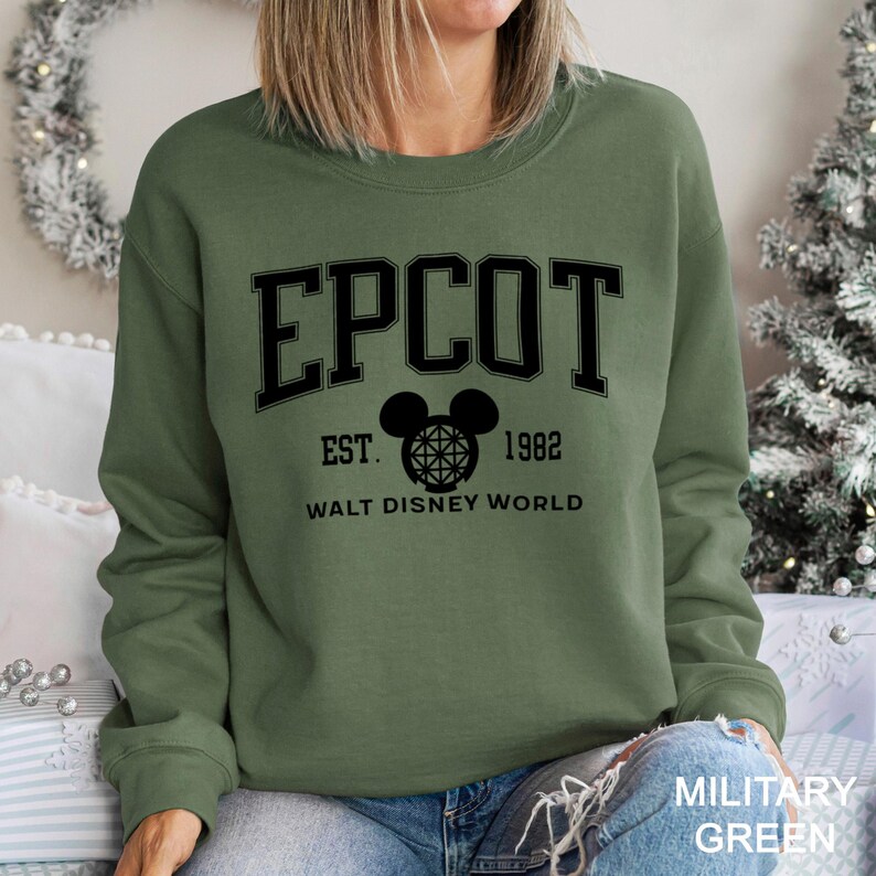 K&ouml;nnte beinhalten: Milit&auml;rgr&uuml;nes Sweatshirt mit dem Wort "EPCOT" in schwarzen Blockbuchstaben. Darunter ein Mickey Mouse-Kopf mit Globus-Design, der Text "EST. 1982" und "WALT DISNEY WORLD". Das Sweatshirt hat einen Rundhalsausschnitt.