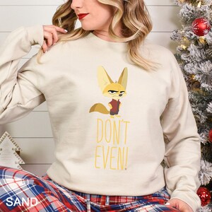 May include: A sand-coloured sweatshirt with a cartoon fennec fox wearing a red vest and an annoyed expression. The yellow text "DON'T EVEN!" is printed below the fox. The sweatshirt has long sleeves.