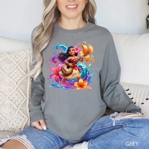 Puede incluir: Una camiseta gris de manga larga con una colorida ilustraci&oacute;n de un personaje de dibujos animados tocando el ukelele, rodeado de flores y salpicaduras de agua. La palabra "GREY" est&aacute; impresa en la esquina inferior derecha de la camiseta.