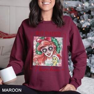 May include: Maroon sweatshirt featuring a graphic of Ariel from The Little Mermaid wearing sunglasses and a Santa hat with the text "BUT DADDY I love him". The design is framed in a red rectangle. The word "MAROON" is printed at the bottom.