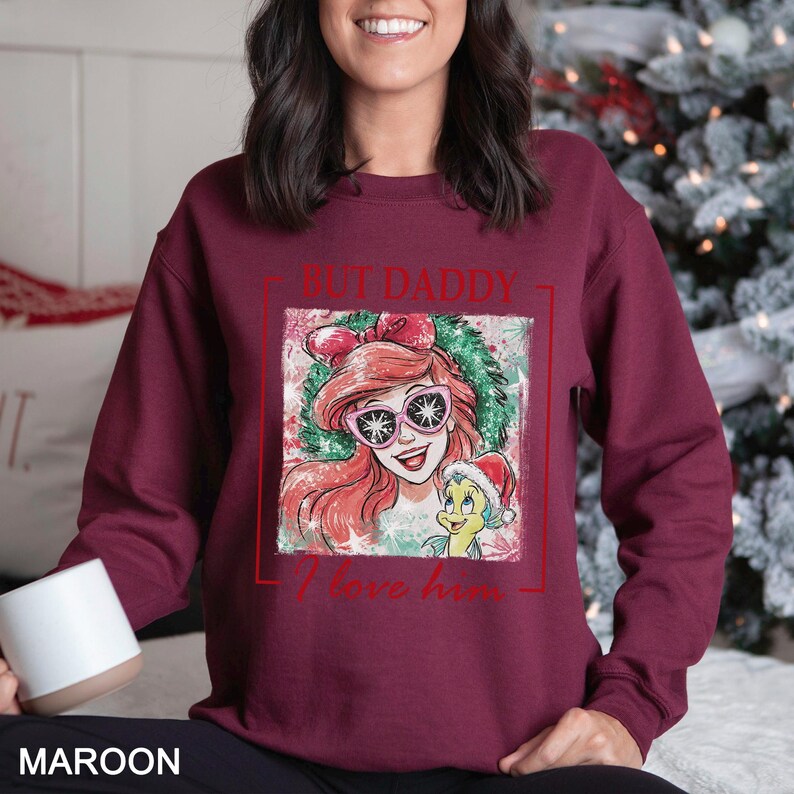 May include: Maroon sweatshirt featuring a graphic of Ariel from The Little Mermaid wearing sunglasses and a Santa hat with the text "BUT DADDY I love him". The design is framed in a red rectangle. The word "MAROON" is printed at the bottom.