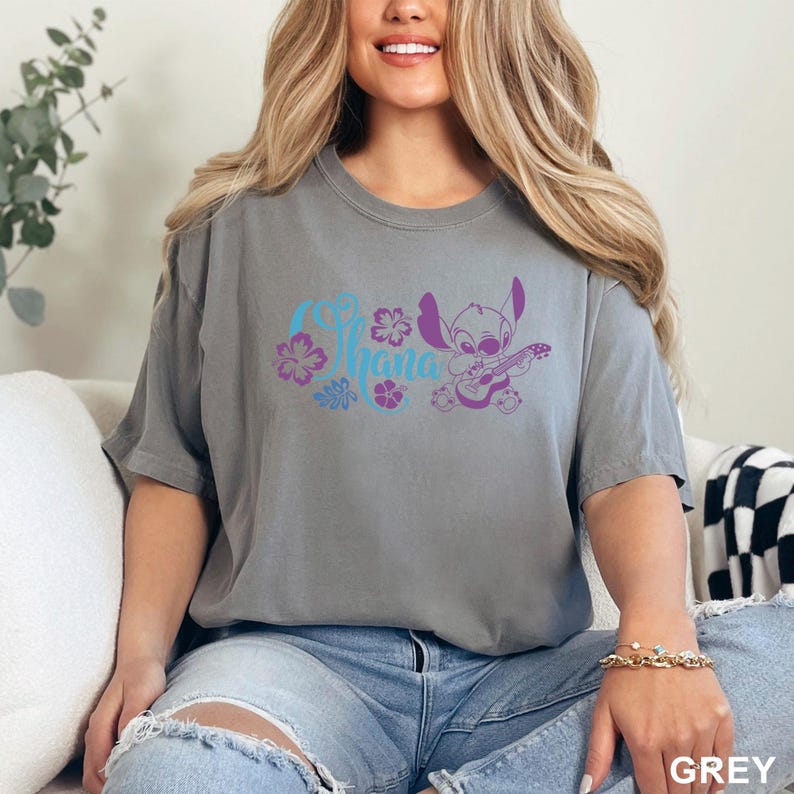 Puede incluir: Camiseta gris con la palabra "Ohana" en escritura azul y morada, acompa&ntilde;ada de dise&ntilde;os florales y un personaje de dibujos animados tocando la guitarra. La camiseta es de color gris liso.