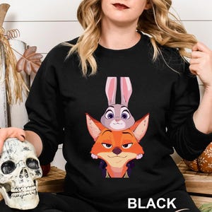 May include: Black crewneck sweatshirt featuring a cartoon image of a fox and a rabbit. The fox is orange and the rabbit is gray with pink ears. The word "BLACK" is printed at the bottom of the sweatshirt.
