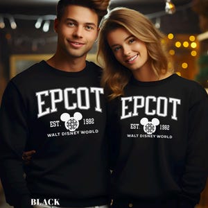 K&ouml;nnte beinhalten: Schwarze Sweatshirts mit dem Wort "EPCOT" in wei&szlig;er Schrift im College-Stil. Darunter steht der Text "EST. 1982 WALT DISNEY WORLD" in Wei&szlig;, mit einer Mickey-Mouse-Kopf-Grafik. Das Wort "BLACK" steht unten.