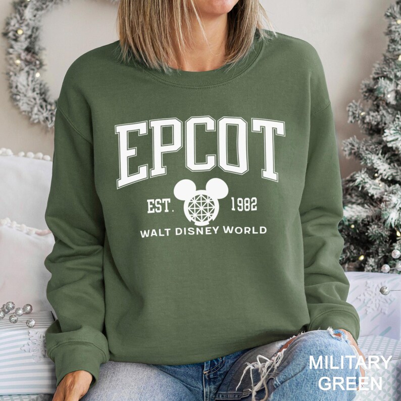 K&ouml;nnte beinhalten: Milit&auml;rgr&uuml;nes Sweatshirt mit dem Wort "EPCOT" in wei&szlig;en Blockbuchstaben. Darunter befindet sich ein Mickey-Mouse-Kopf-Logo mit dem Text "EST. 1982 WALT DISNEY WORLD". Das Sweatshirt ist einfarbig und hat lange &Auml;rmel.