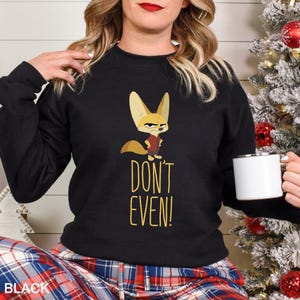 May include: Black sweatshirt with a cartoon fennec fox wearing a red vest. The text "DON'T EVEN!" is below the fox in yellow. The sweatshirt is worn with plaid trousers. The image also shows a white mug.