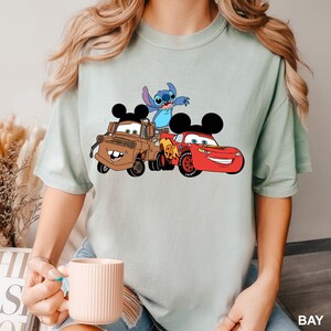 May include: A light blue t-shirt with a graphic print of Mickey Mouse, Stitch, and Lightning McQueen from Disney's Cars.