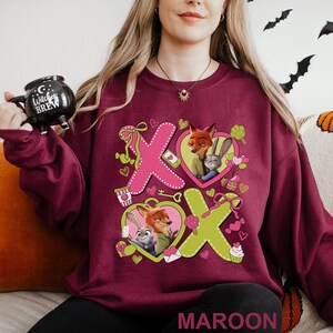 May include: Maroon sweatshirt featuring a graphic of the characters Nick Wilde and Judy Hopps from Zootopia, with the letters X and O in pink and green. The design includes hearts, bows, and other Valentine's Day themed elements.