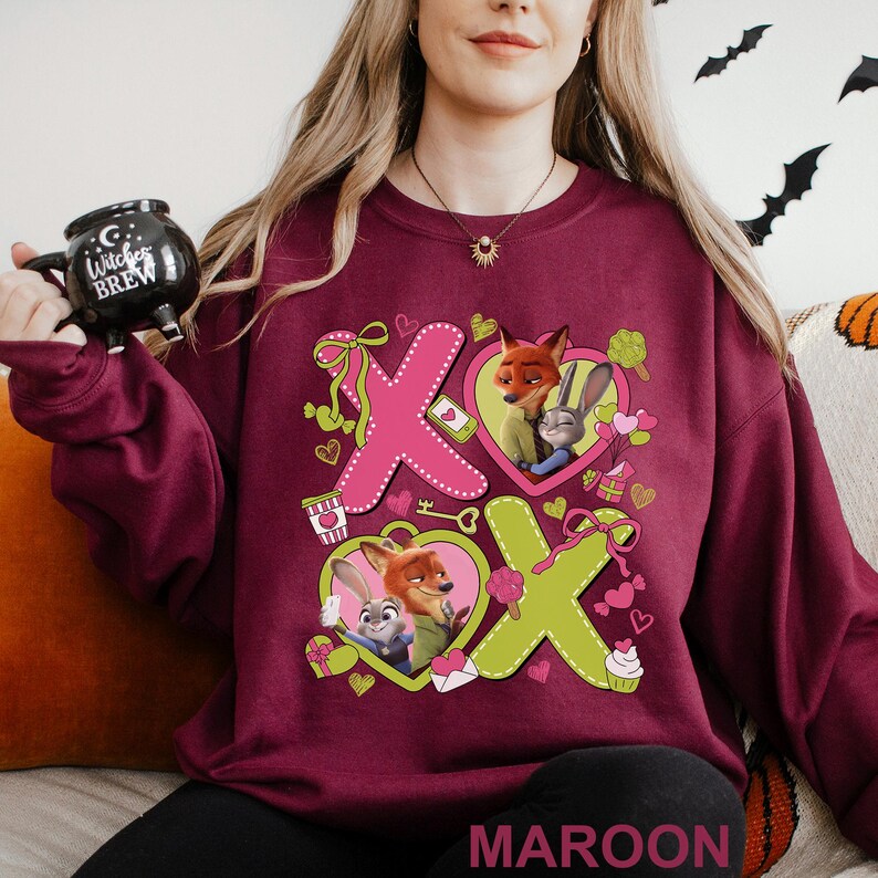 May include: Maroon sweatshirt featuring a graphic of the characters Nick Wilde and Judy Hopps from Zootopia, with the letters X and O in pink and green. The design includes hearts, bows, and other Valentine's Day themed elements.