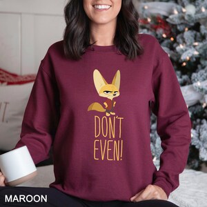 May include: Maroon sweatshirt with a cartoon fox illustration and the text "DON'T EVEN!". The fox has an angry expression and wears a red vest. The text and fox are in yellow. The sweatshirt is a solid maroon colour.