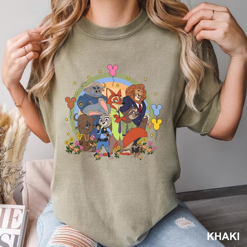 May include: Khaki-colored t-shirt featuring a colorful graphic of characters from the animated movie Zootopia. The design includes Judy Hopps, Nick Wilde, and other characters, surrounded by flowers and balloons. The word "KHAKI" is printed at the bottom.