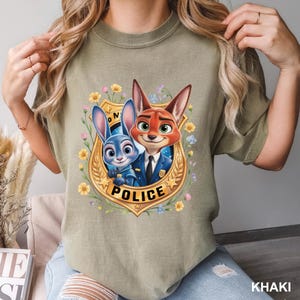 K&ouml;nnte beinhalten: Khaki-farbenes T-Shirt mit einem Cartoon-Bild von Judy Hopps und Nick Wilde aus Zootopia in einem goldenen Polizeiausweis. Die Auszeichnung ist von bunten Blumen umgeben. Das Wort "POLICE" ist auf dem Ausweis aufgedruckt.