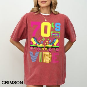 May include: A crimson red t-shirt with a graphic design featuring the text "70's Vibe" in colorful, outlined letters. The design also includes a pair of yellow roller skates, a peace sign, musical notes, and a cassette tape.