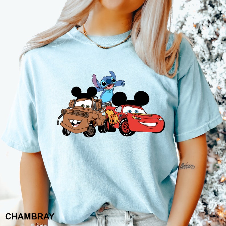 May include: A light blue t-shirt with a graphic print of Stitch, Mater, and Lightning McQueen from Disney. The characters are in a cartoon style and are wearing Mickey Mouse ears.