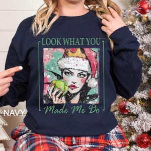May include: Navy blue sweatshirt with a graphic of a woman wearing a Santa hat and holding a green apple. The text "LOOK WHAT YOU MADE ME DO" is printed on the sweatshirt. The word "NAVY" is printed on the left side of the sweatshirt.
