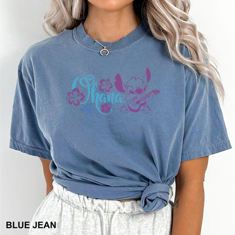 Puede incluir: Camiseta azul vaquero con la palabra "Ohana" en escritura turquesa y detalles florales rosas. Un personaje de dibujos animados morado tocando la guitarra est&aacute; a la derecha de la palabra. La camiseta tiene mangas cortas y cuello redondo.