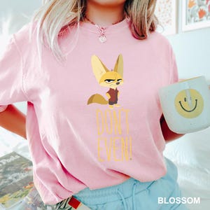 May include: A light pink t-shirt featuring a cartoon fennec fox with a stern expression. The text "DON'T EVEN!" is printed below the fox in yellow. A white mug with a yellow smiley face is held in the hand.