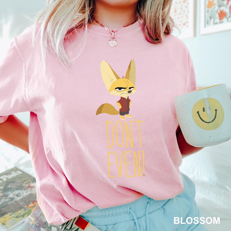 May include: A light pink t-shirt featuring a cartoon fennec fox with a stern expression. The text "DON'T EVEN!" is printed below the fox in yellow. A white mug with a yellow smiley face is held in the hand.