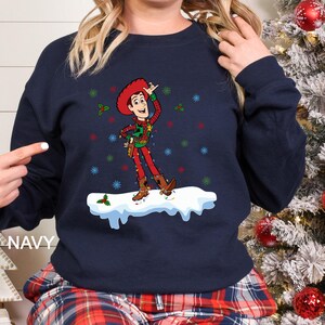 Puede incluir: Sudadera azul marino con un dise&ntilde;o navide&ntilde;o de un vaquero de dibujos animados. El vaquero lleva un atuendo rojo y verde con luces navide&ntilde;as y est&aacute; sobre una superficie nevada. La palabra "NAVY" est&aacute; impresa en el lado izquierdo.