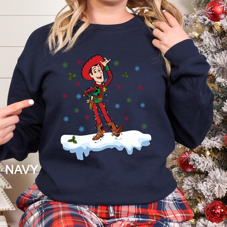 Puede incluir: Sudadera azul marino con un dise&ntilde;o navide&ntilde;o de un vaquero de dibujos animados. El vaquero lleva un atuendo rojo y verde con luces navide&ntilde;as y est&aacute; sobre una superficie nevada. La palabra "NAVY" est&aacute; impresa en el lado izquierdo.