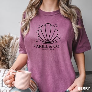 May include: A mauve colored t-shirt with a black graphic of a seashell and the text "Ariel & Co. Since 1989".