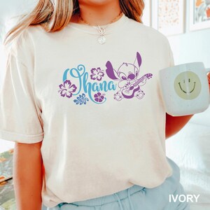 Puede incluir: Camiseta color marfil con la palabra "Ohana" en escritura azul, detalles florales morados y un personaje de dibujos animados morado tocando la guitarra. Tambi&eacute;n se ve una taza blanca con una carita sonriente verde.