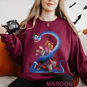 May include: Maroon sweatshirt featuring a cartoon graphic of a blue snake wrapped around a fox and a rabbit. The word "MAROON" is printed at the bottom. The person is also holding a black mug with the words "Witches Brew".
