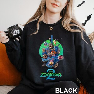 May include: Black crewneck sweatshirt featuring a colorful graphic of characters from the Disney movie "Zootopia 2" inside a green circle. The text "ZOOTOPIA 2" is below the graphic in teal. The word "BLACK" is at the bottom.