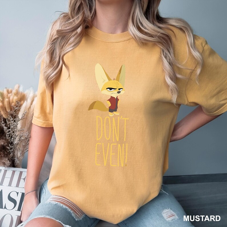 May include: Mustard-colored t-shirt featuring a cartoon fox with a stern expression, wearing a red vest. The text "DON'T EVEN!" is printed below the fox in yellow. The shirt is a relaxed fit.