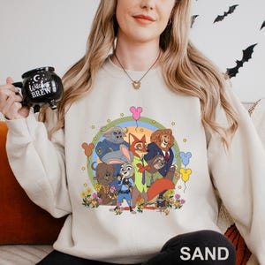 May include: A sand-coloured sweatshirt with a colourful cartoon graphic of animal characters. The graphic includes a rainbow, flowers, and balloons. The word "SAND" is printed at the bottom of the sweatshirt.