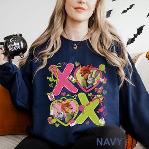 May include: Navy blue sweatshirt featuring a colorful graphic of the characters Nick Wilde and Judy Hopps from Zootopia, with the letters X and O. The design includes hearts, cupcakes, and other decorative elements. The word "NAVY" is printed at the bottom.