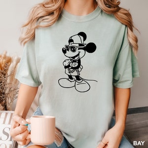 Może przedstawiać: Jasno błękitny t-shirt z czarno-białym nadrukiem przedstawiającym Myszki Miki w okularach przeciwsłonecznych i czapce z daszkiem.