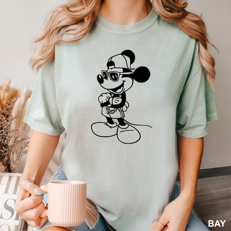 Może przedstawiać: Jasno błękitny t-shirt z czarno-białym nadrukiem przedstawiającym Myszki Miki w okularach przeciwsłonecznych i czapce z daszkiem.