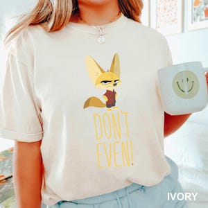 May include: Ivory-colored t-shirt featuring a cartoon fennec fox with a stern expression, wearing a maroon vest. Below the fox, the words "DON'T EVEN!" are printed in yellow. A white mug with a smiley face is visible.