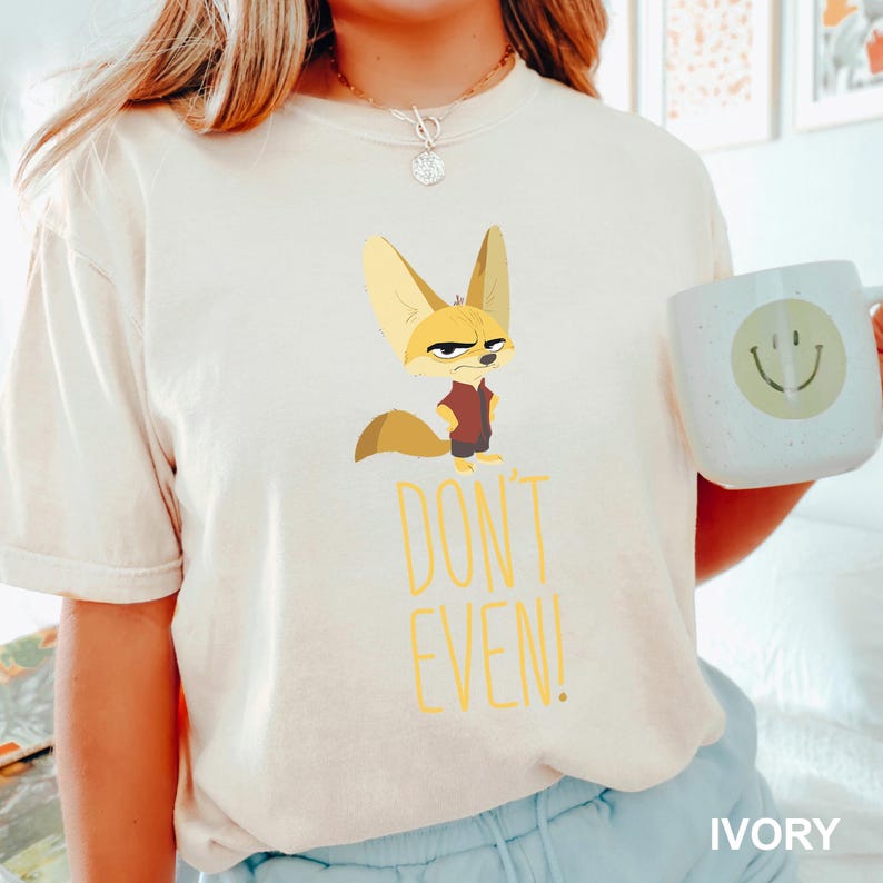 May include: Ivory-colored t-shirt featuring a cartoon fennec fox with a stern expression, wearing a maroon vest. Below the fox, the words "DON'T EVEN!" are printed in yellow. A white mug with a smiley face is visible.