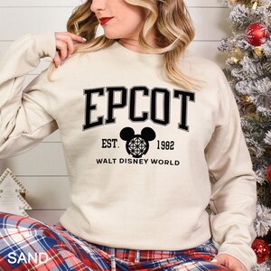K&ouml;nnte beinhalten: Ein sandfarbener Pullover mit dem Wort "EPCOT" in schwarzen Blockbuchstaben. Darunter ein schwarzer Mickey Mouse-Kopf mit geometrischem Design, der Text "EST. 1982" und "WALT DISNEY WORLD". Der Pullover hat lange &Auml;rmel.