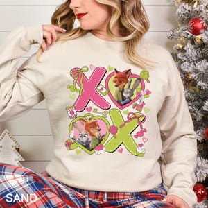 May include: A sand-colored sweatshirt featuring a pink and green design with the letters "XO" and images of cartoon characters in heart shapes. The sweatshirt has long sleeves and a crew neck.