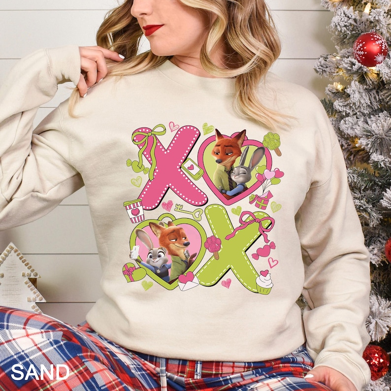May include: A sand-colored sweatshirt featuring a pink and green design with the letters "XO" and images of cartoon characters in heart shapes. The sweatshirt has long sleeves and a crew neck.